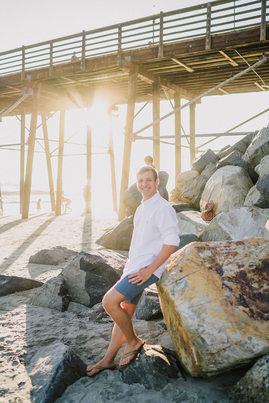 Smiling young man in white shirt leaning on rocks under pier during golden hour at beach.