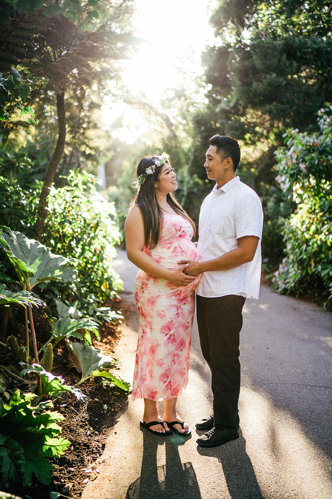 Pregnant couple holding baby bump in a sunlit garden path, woman in floral dress with flower crown, man in white shirt.