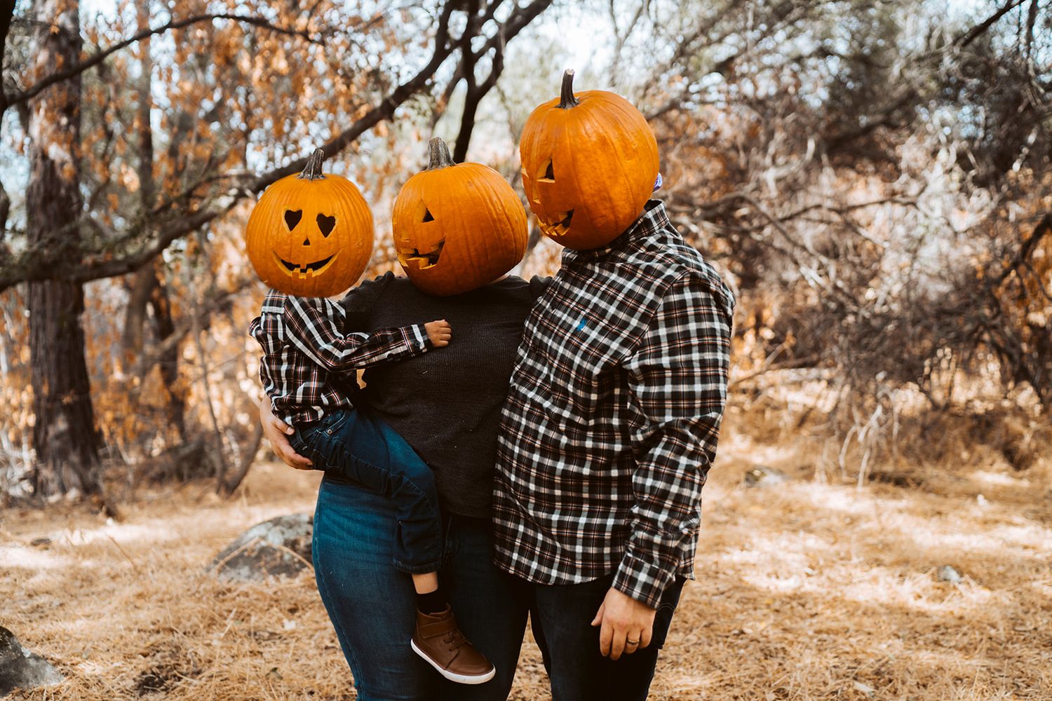 Couple holds bright orange pumpkins in a rustic outdoor autumn setting with fall foliage in background.
