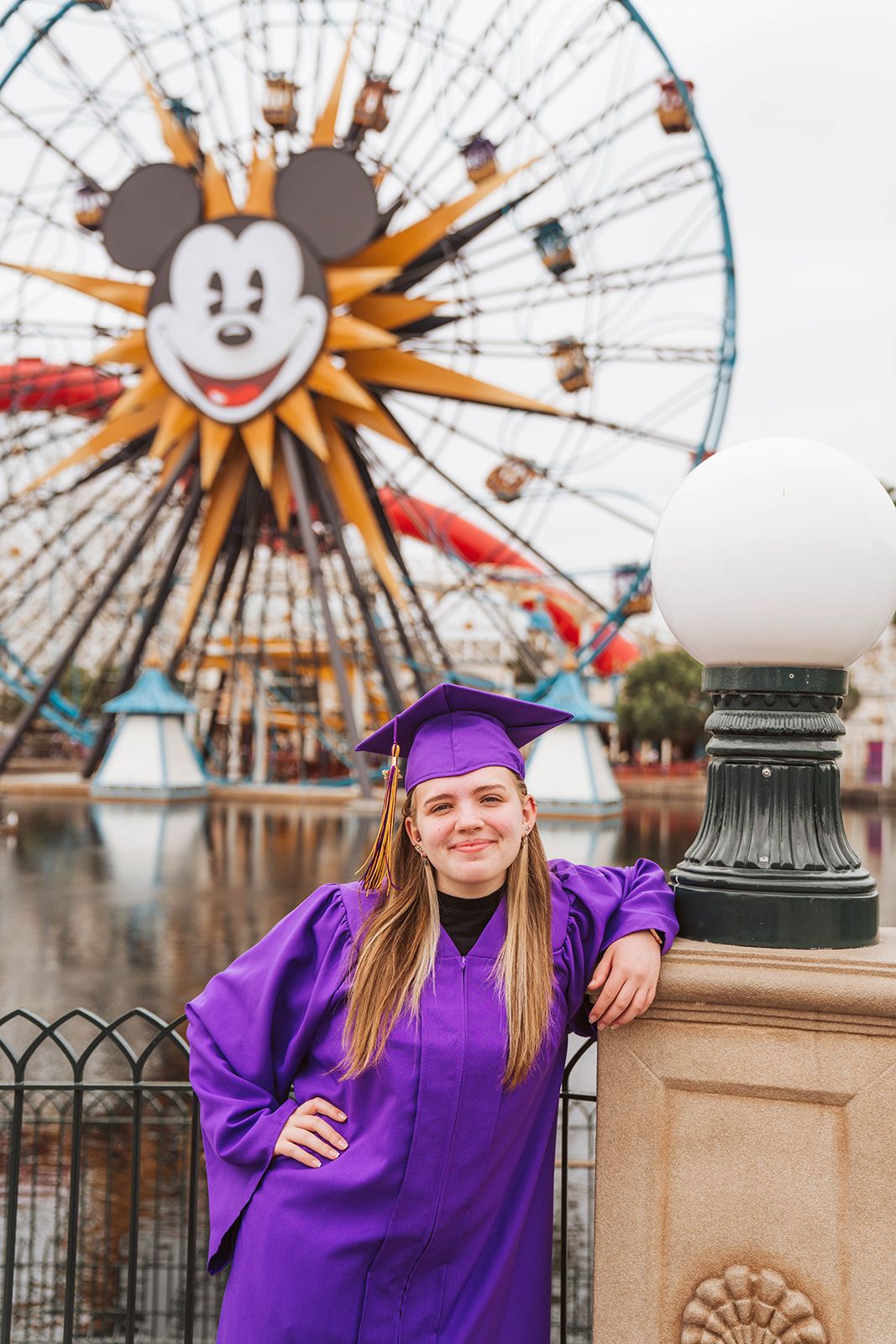 Graduate in purple cap and gown poses in front of Mickey Mouse Ferris wheel at Disney California Adventure.