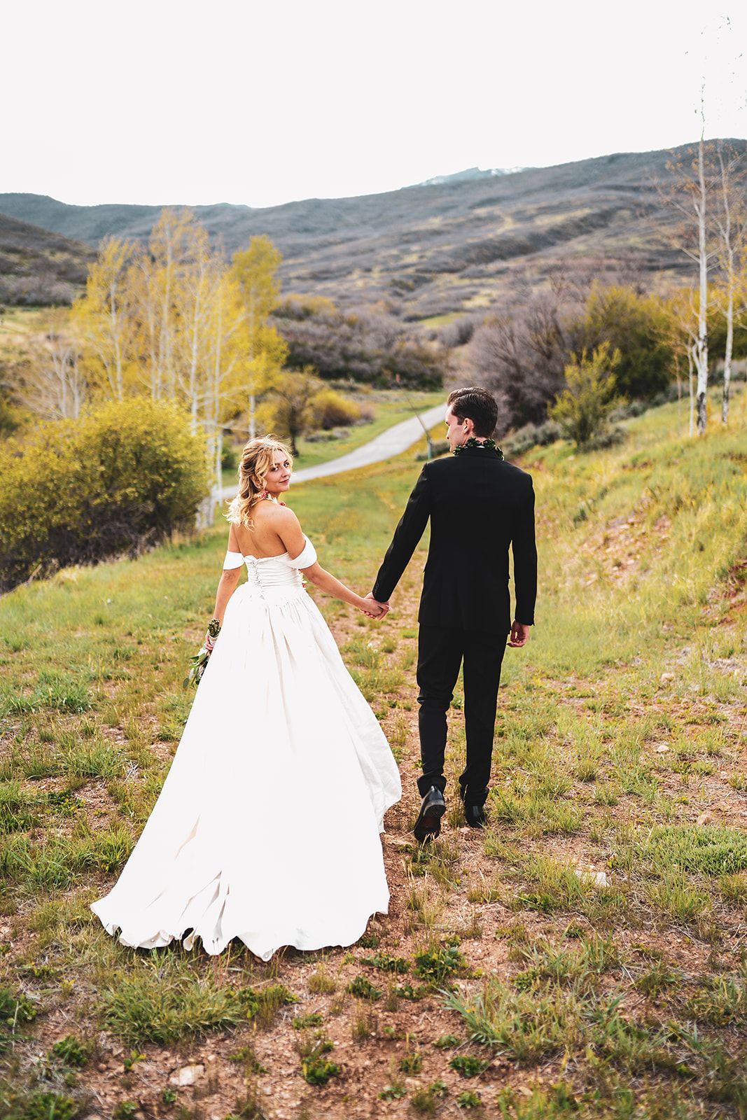 Bride in white gown and groom in black suit walk hand-in-hand through a lush green mountain landscape.