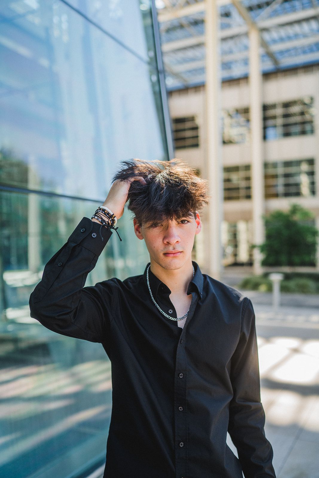 Young man in black shirt runs hand through messy dark hair, posing outdoors near modern glass building in sunlight.