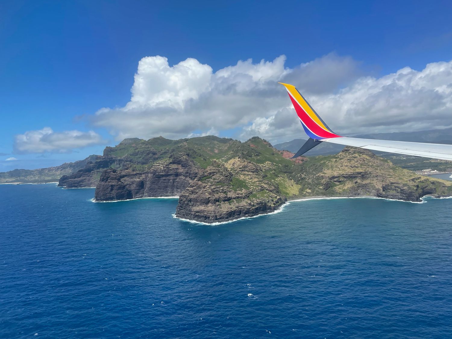 An airplane wing soars over the dramatic cliffs and turquoise waters of the Hawaiian coastline on a sunny day.