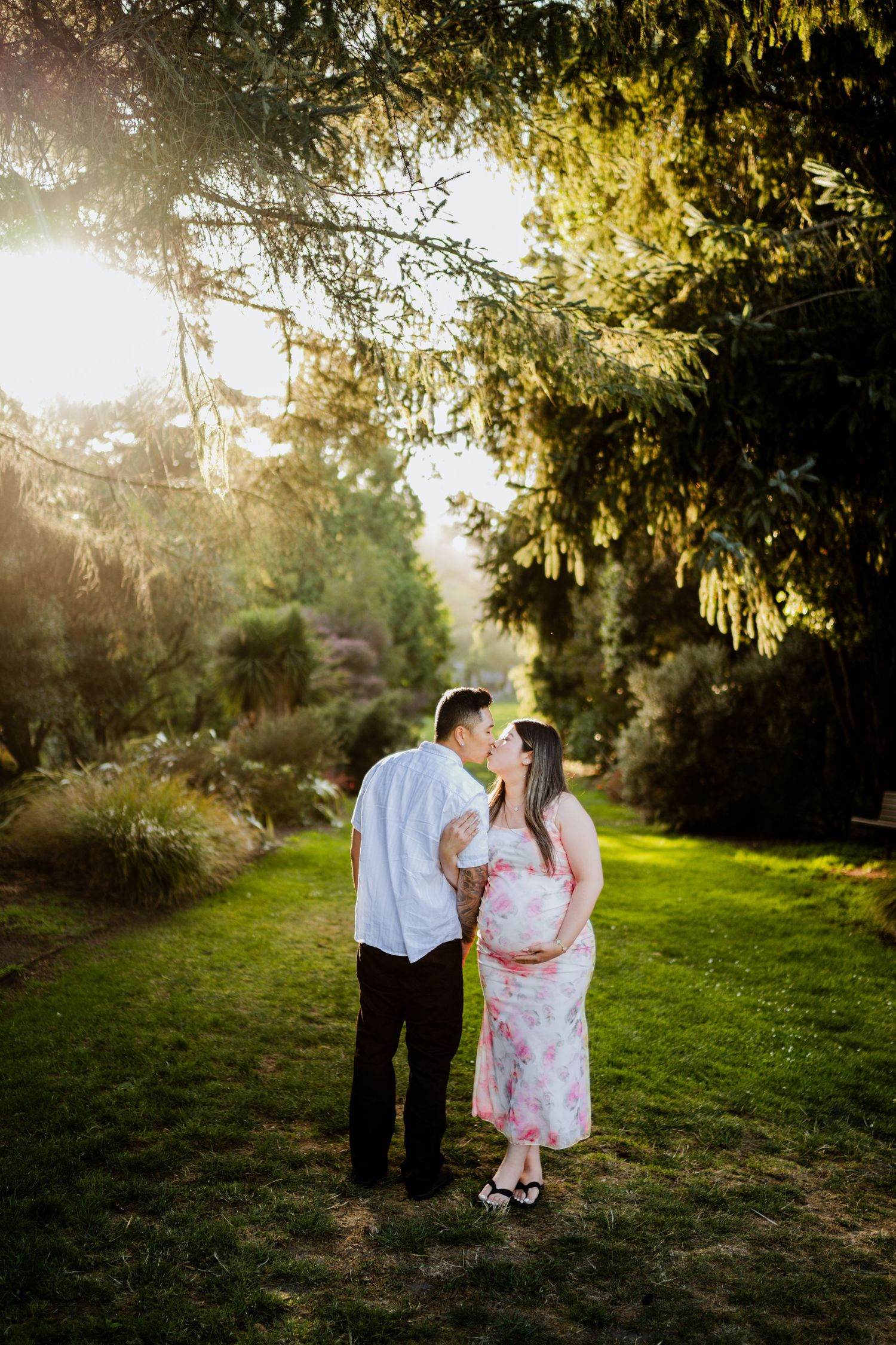 Couple sharing a tender moment in a sunlit garden, woman in floral dress showing pregnancy bump.