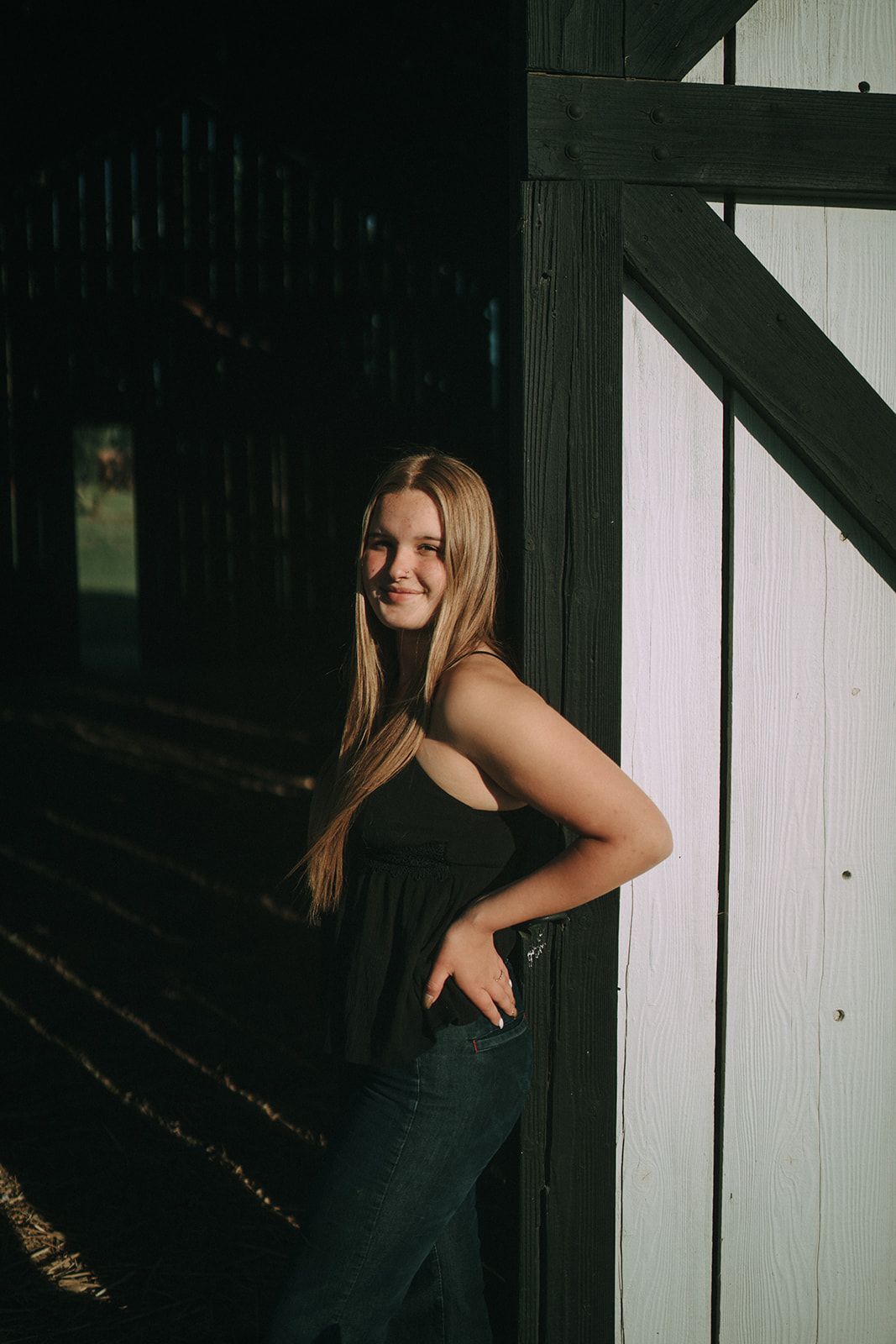 Young blonde woman in black top and jeans posing by barn door in dramatic light and shadow.