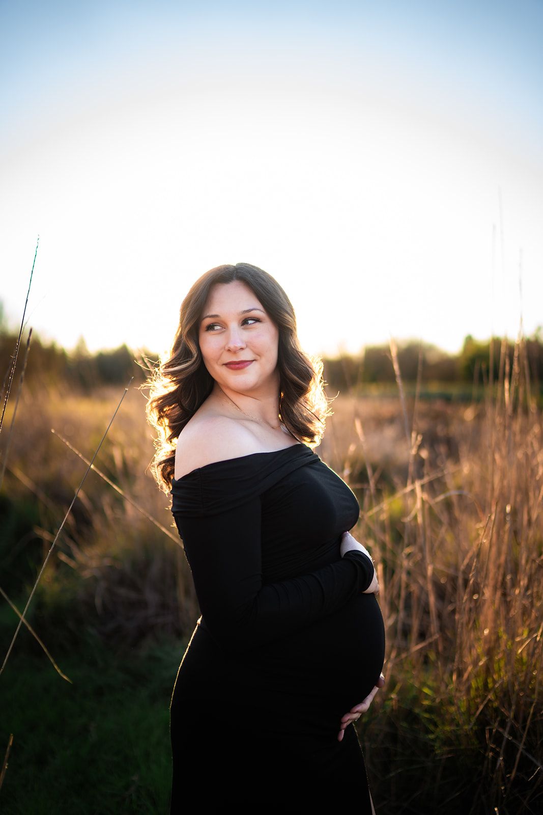 Maternity portrait of woman in black dress during golden hour in tall grass field.