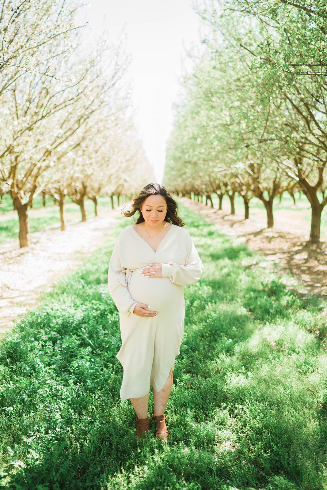 Pregnant woman in white dress cradling baby bump while walking through a sunlit orchard with green grass.