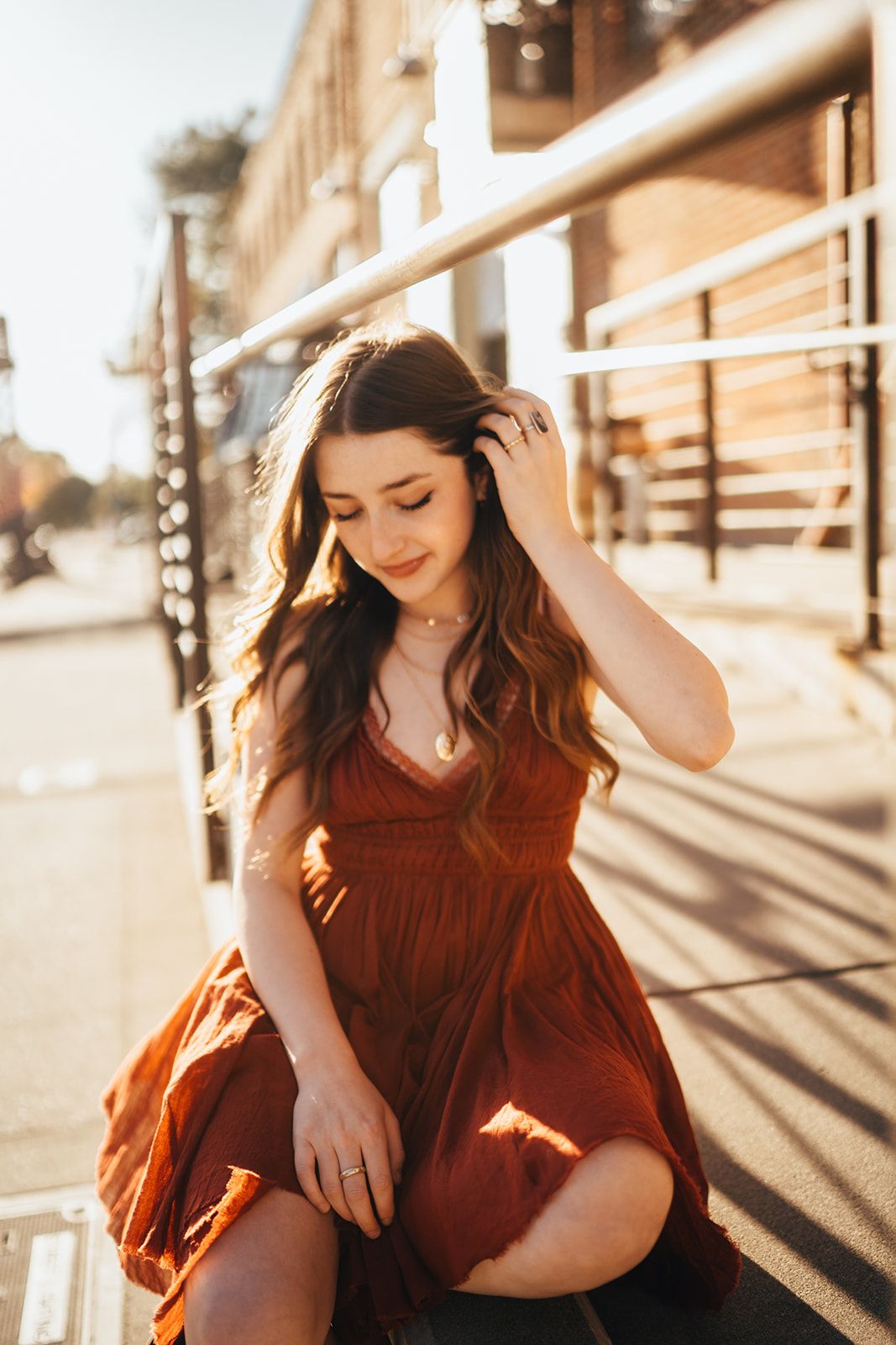 Young woman in rust-colored dress sitting on steps in golden hour sunlight, touching her hair.