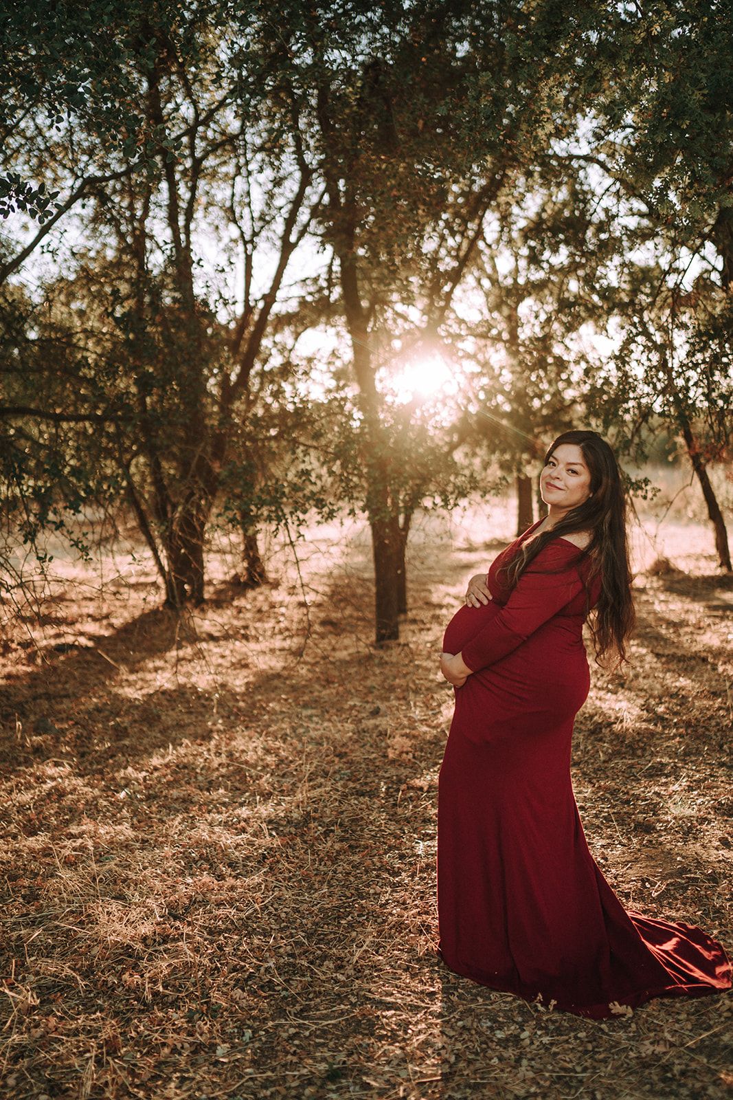 Pregnant woman in red gown standing in sunlit forest at golden hour, cradling baby bump with crossed arms.