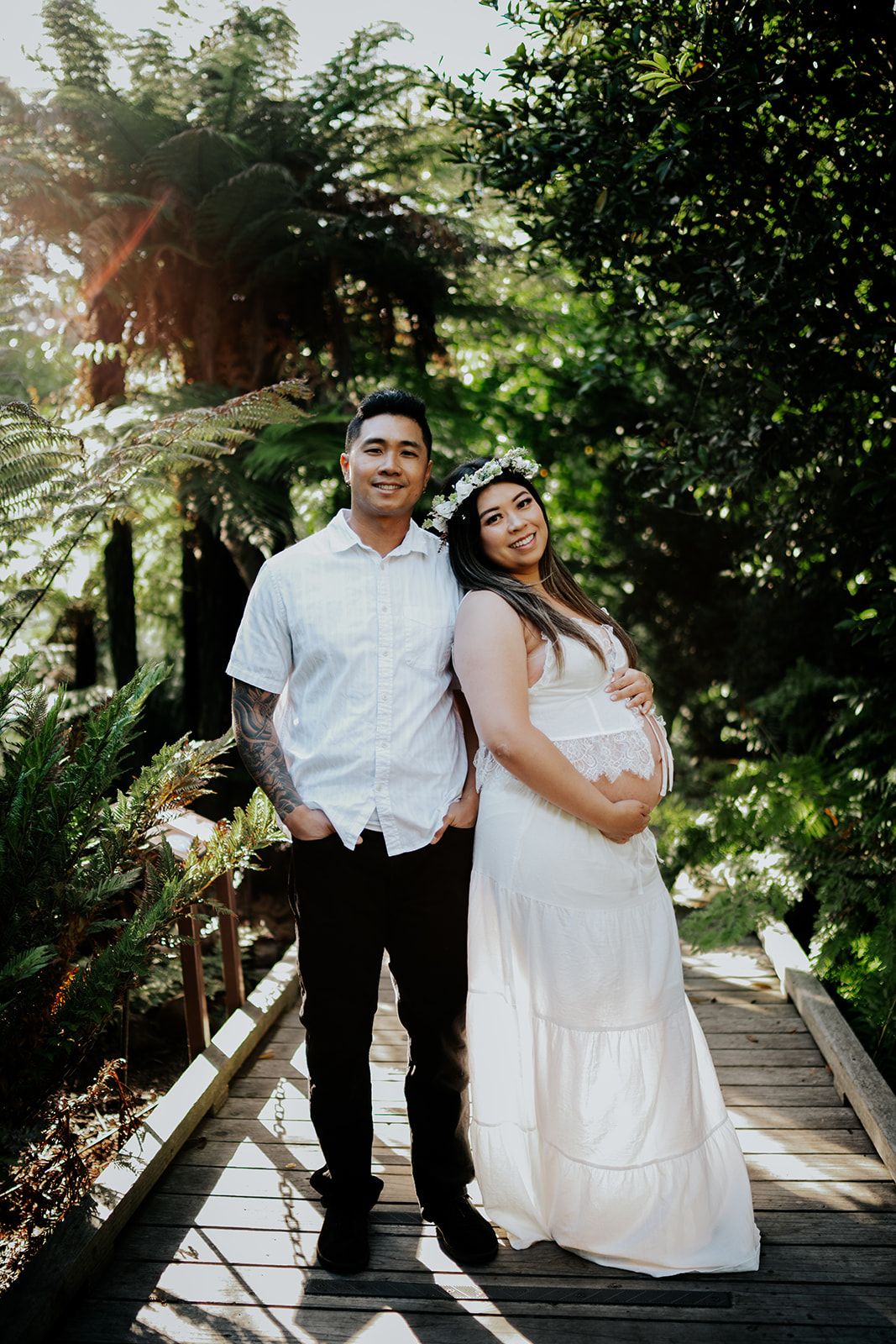Couple in white outfits on wooden bridge surrounded by lush greenery, pregnant woman wearing floral crown during maternity photoshoot.