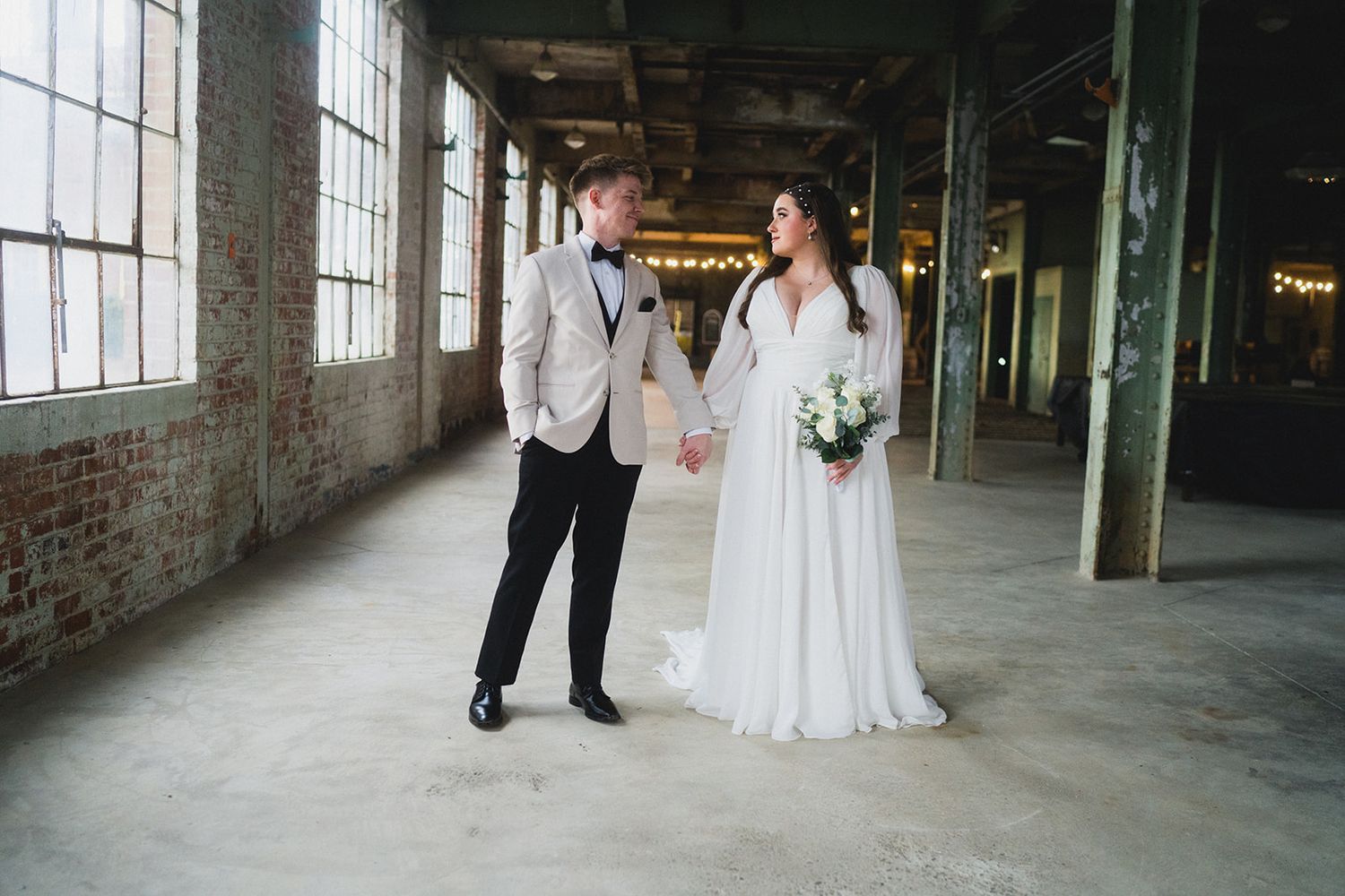 Wedding couple poses in an industrial warehouse setting with large windows and concrete floors.