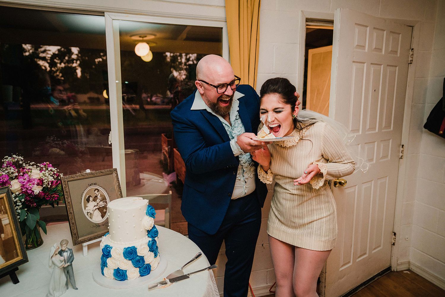Playful moment while feeding wedding cake in front of floral decorations and blue-tiered dessert.