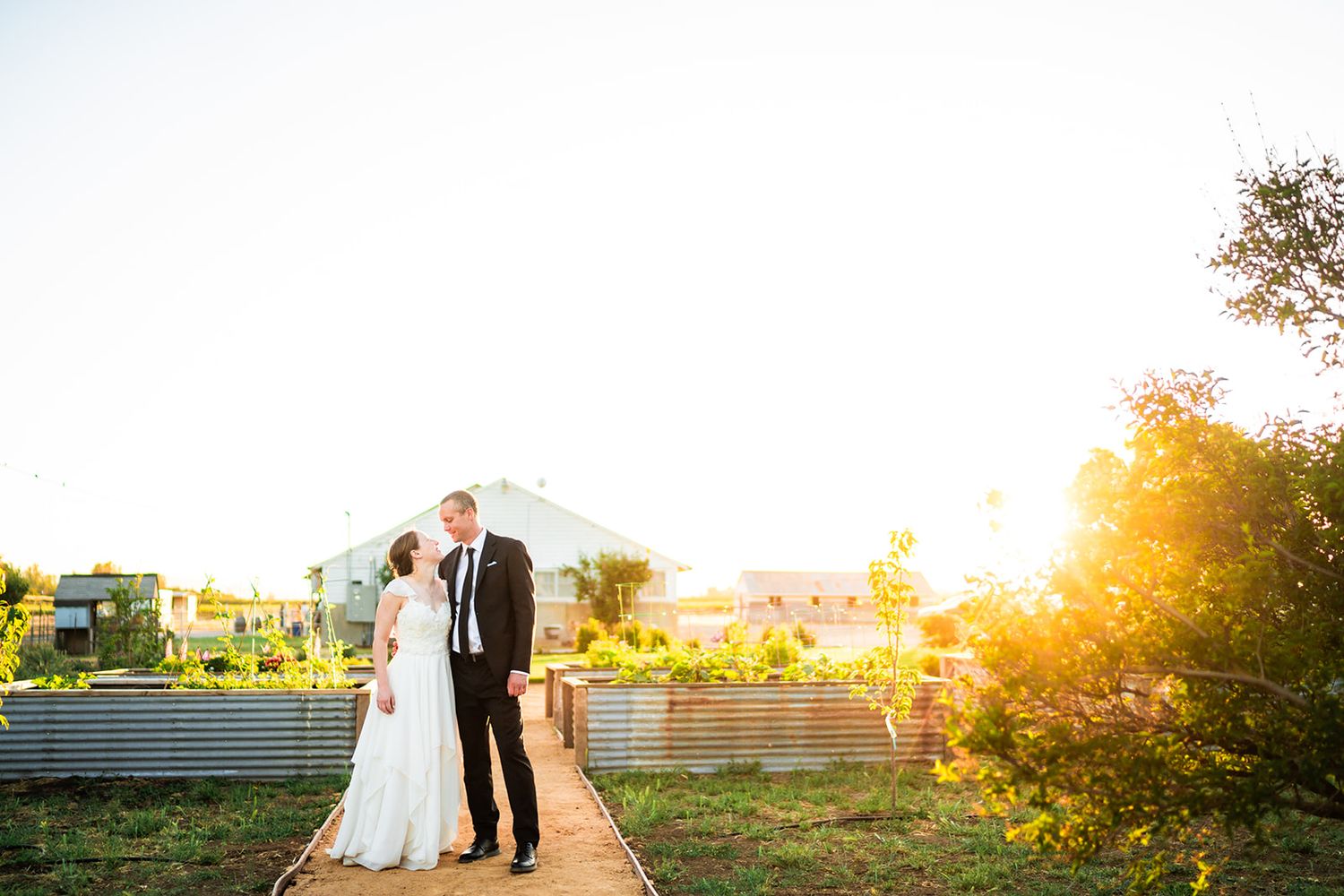 Bride and groom share a romantic moment on a garden path bathed in golden sunset light at an outdoor wedding venue.