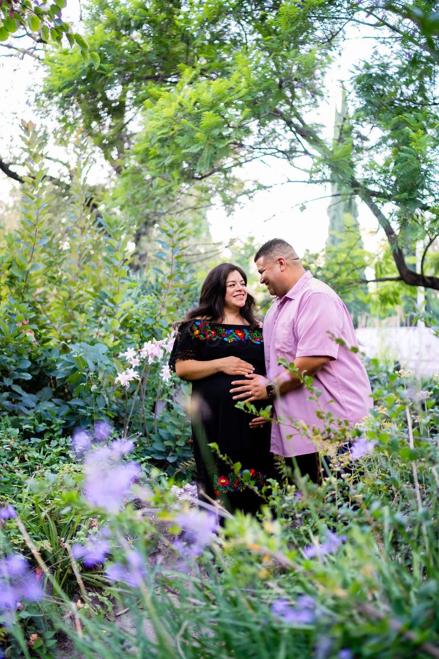 Couple in a lush garden, woman in black embroidered dress with baby bump, man in pink shirt, surrounded by colorful flowers.