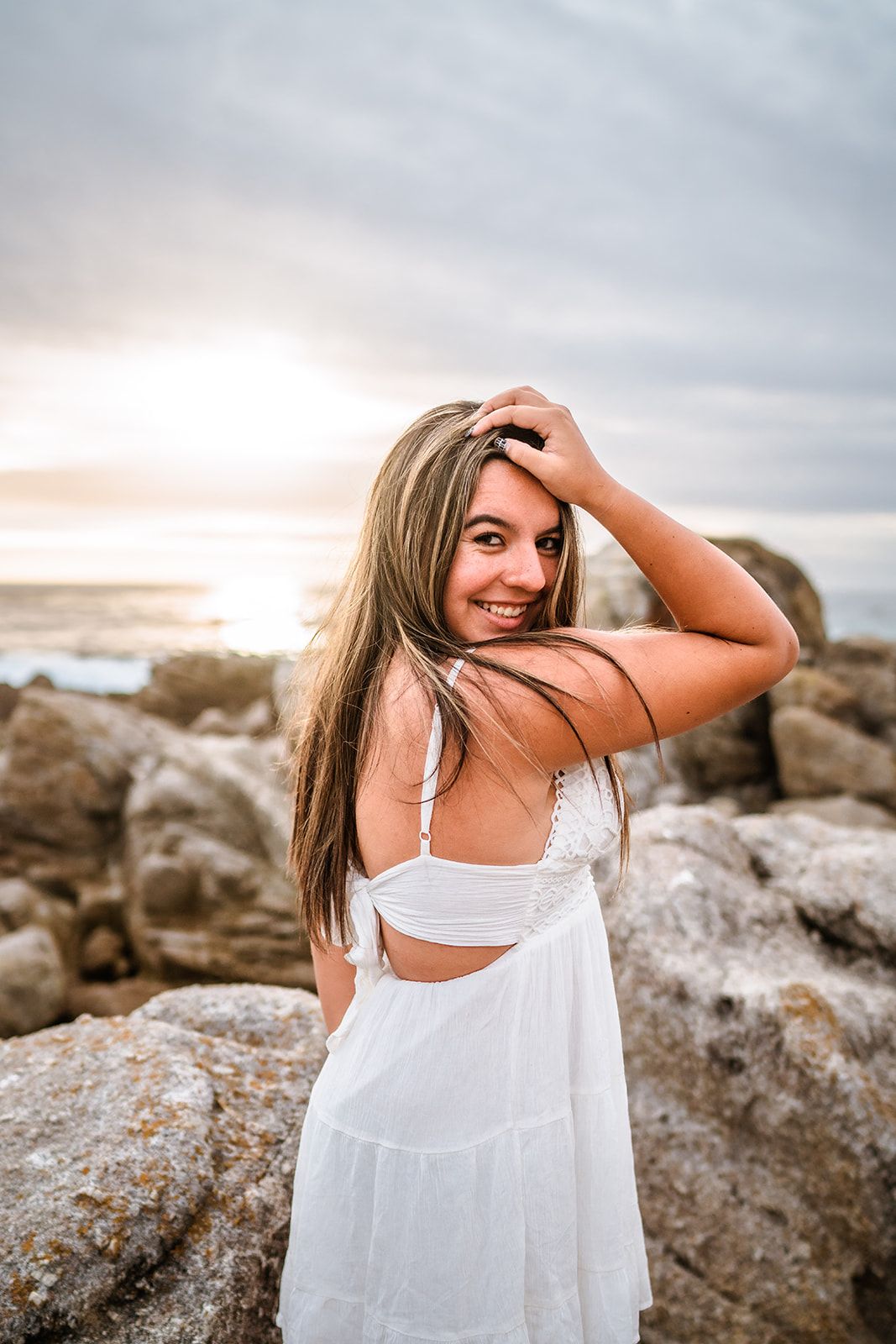 Young woman in white sundress smiling over shoulder at rocky beach during golden sunset.