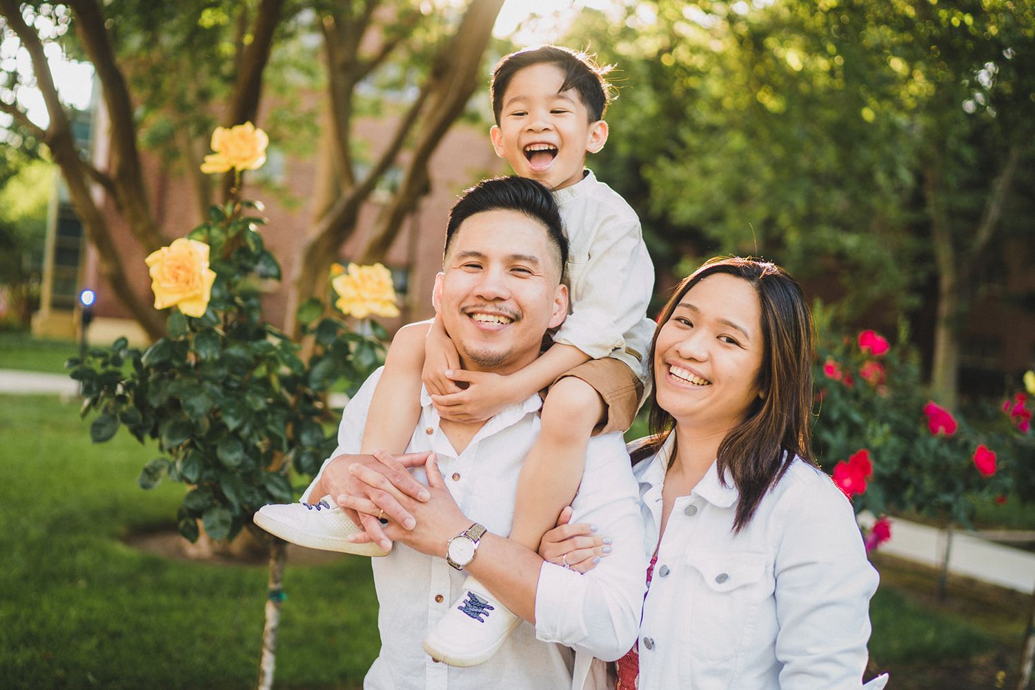 A family in white outfits smiles and embraces together during an outdoor photo session in a sunny garden.