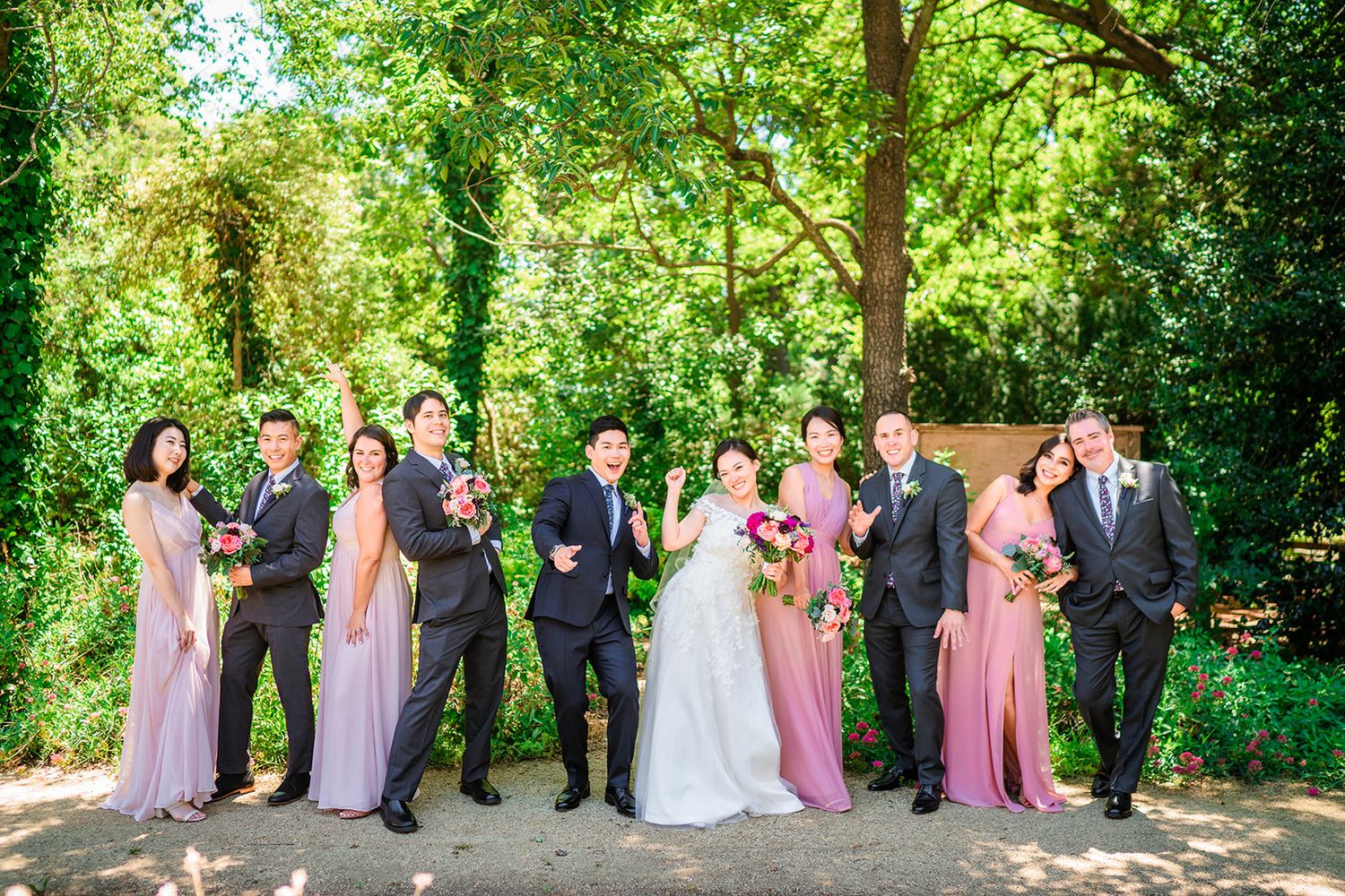 Wedding party poses in a lush green garden wearing pink and gray formal attire.