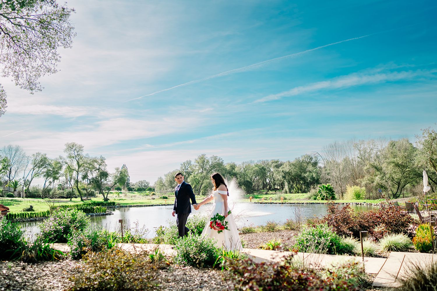 Bride and groom hold hands in a lush garden path beside a serene pond under a bright blue sky.