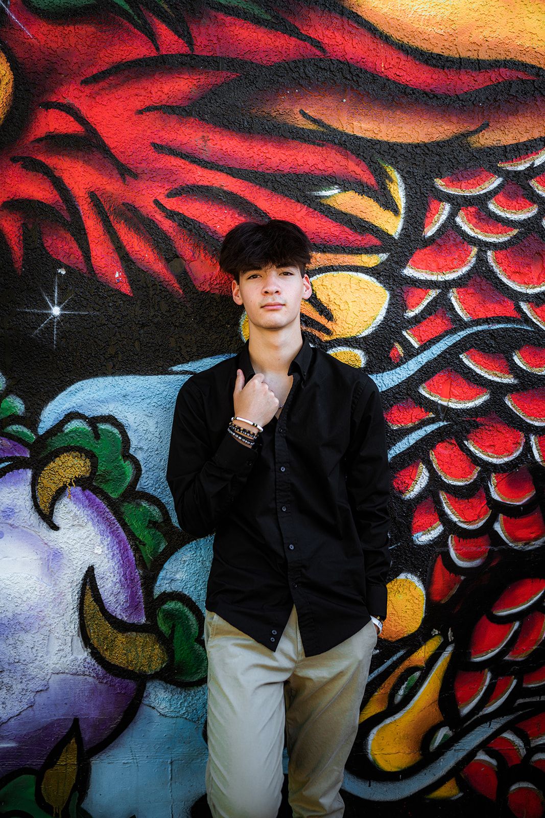 Young man in black shirt poses confidently against a vibrant colorful graffiti mural wall featuring a dragon.