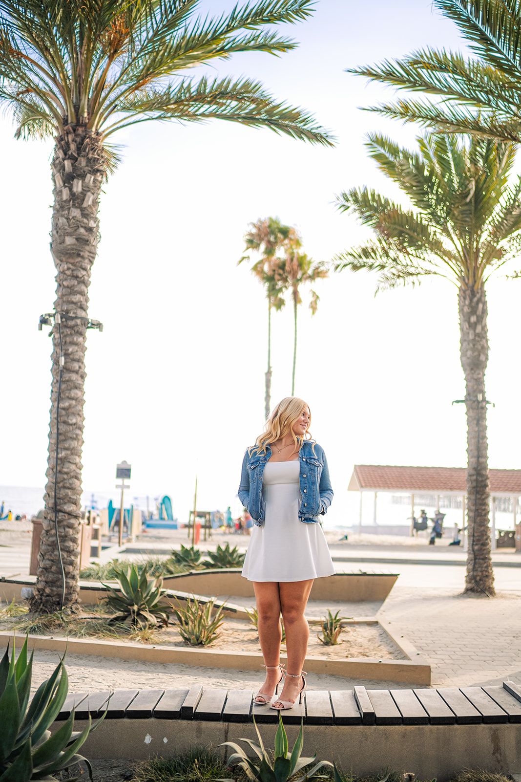 Blonde woman in white dress and denim jacket poses on boardwalk between palm trees near a sunny beach.