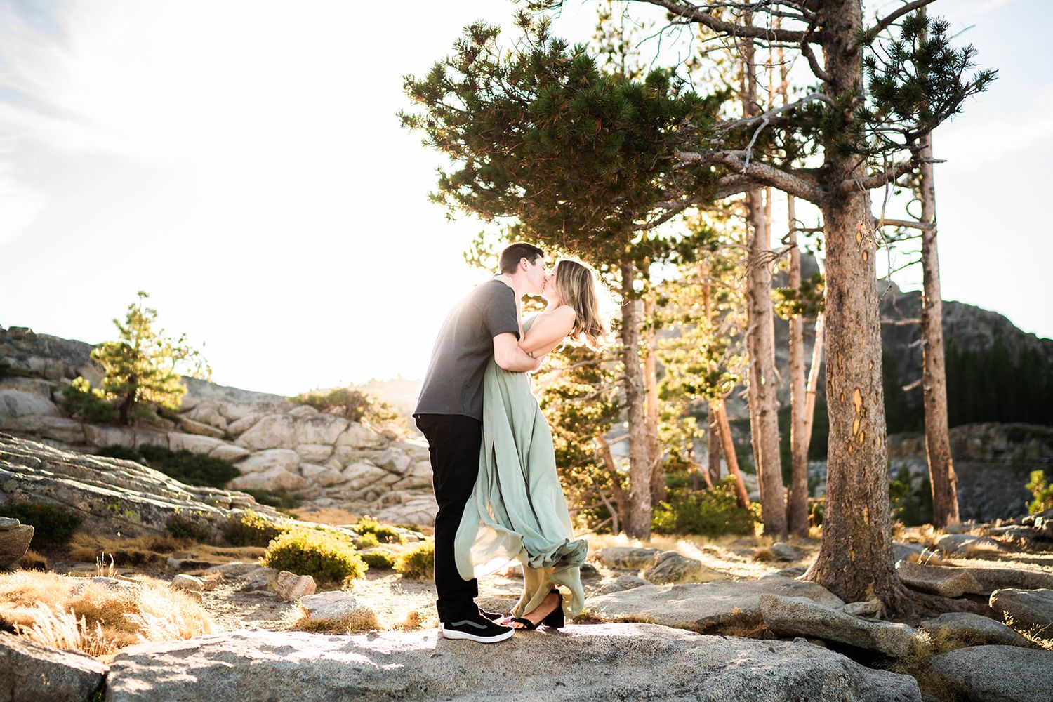 Romantic mountain engagement portrait with couple embracing among pine trees at sunset.