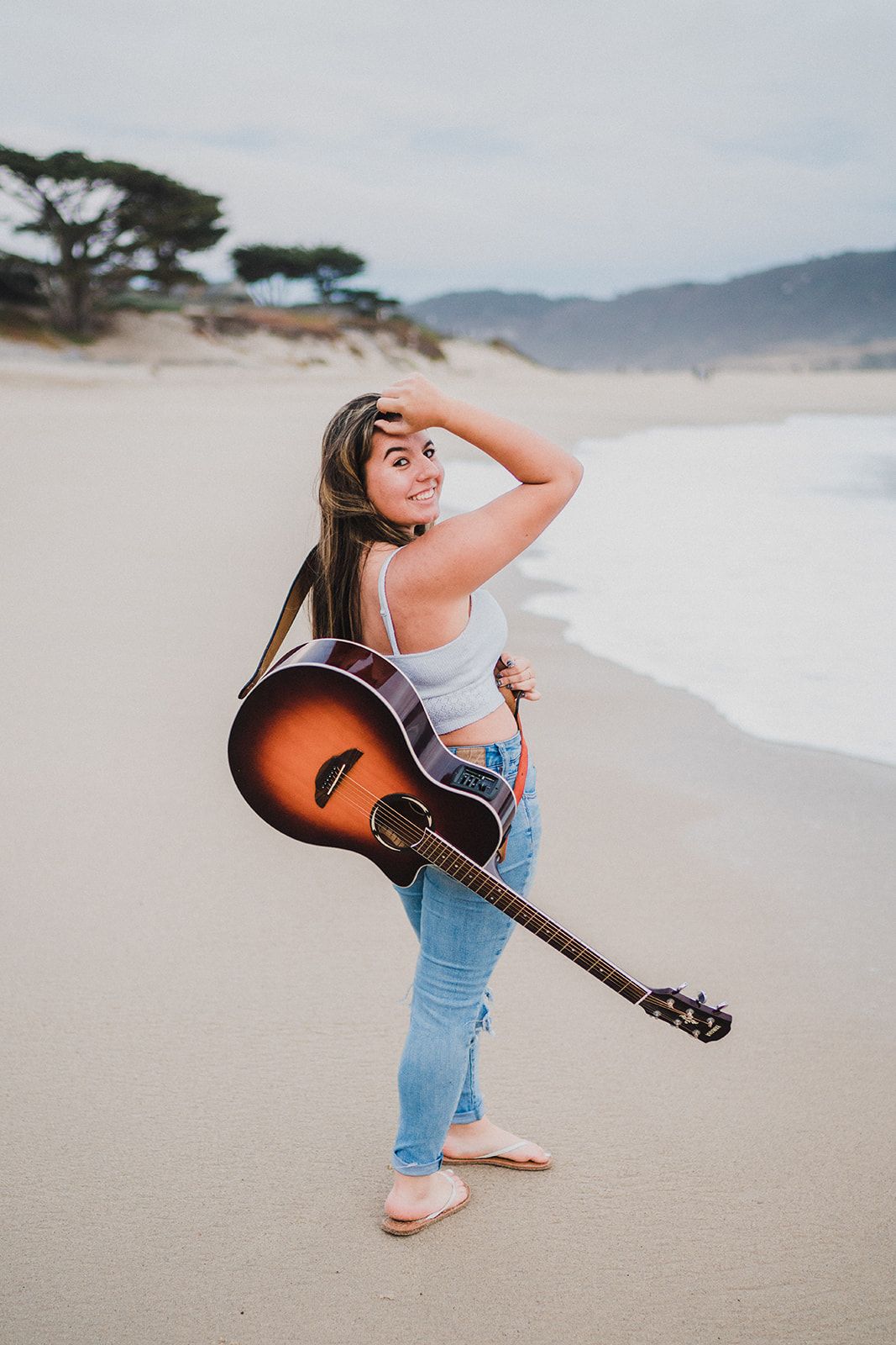 Young woman holding acoustic guitar on sandy beach, wearing white top and jeans, smiling over shoulder.