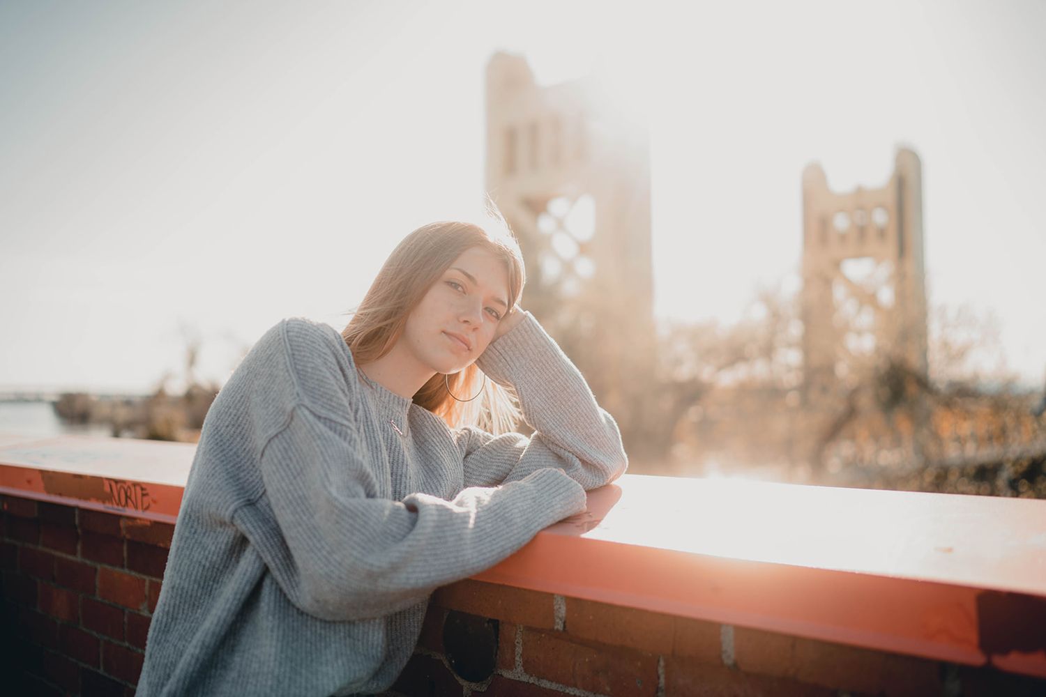 Young woman in gray sweater leans on brick wall with Tower Bridge in golden sunlight background.