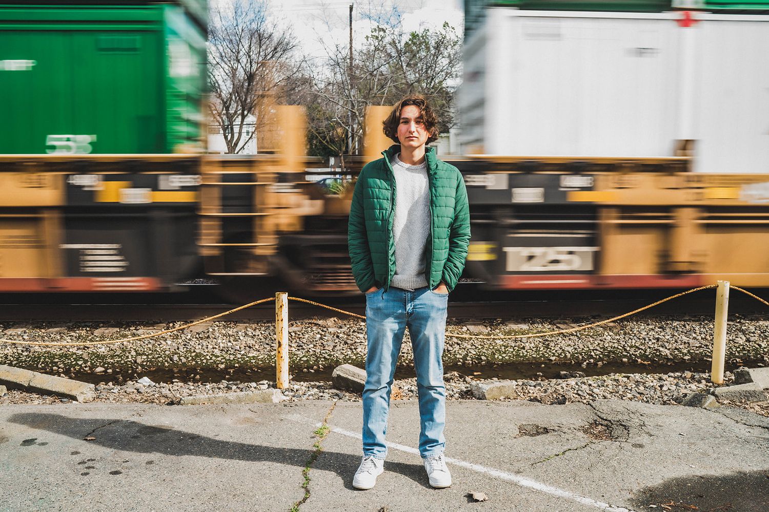 Young man in green jacket and jeans stands confidently in front of a moving freight train on a sunny day.