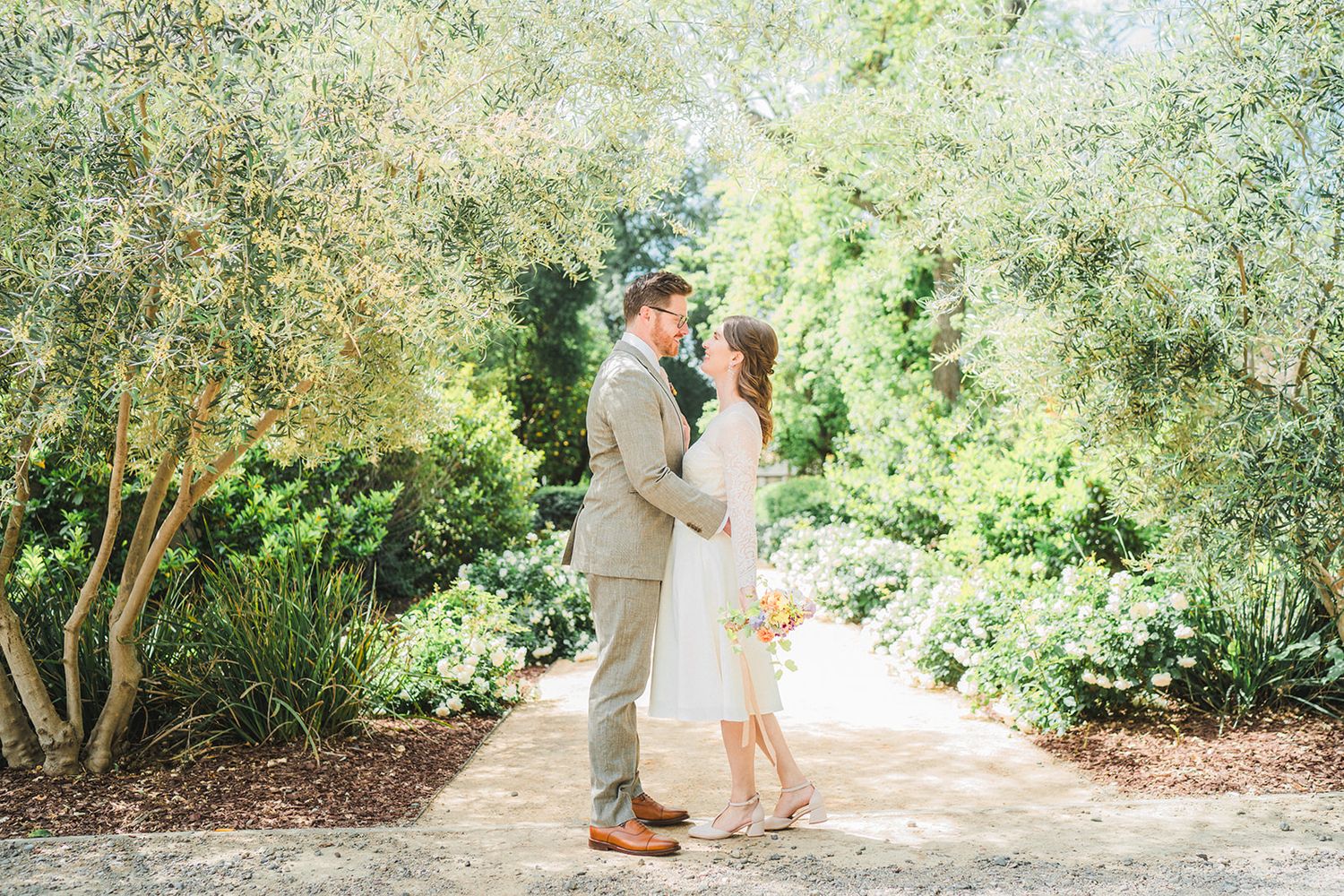 A couple shares a romantic moment on a garden path surrounded by lush greenery and flowering shrubs in natural light.