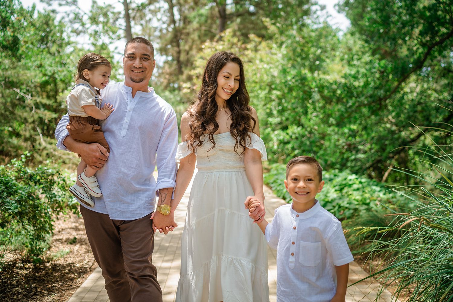 A family walks together on a tree-lined path during a sunny outdoor photo session.