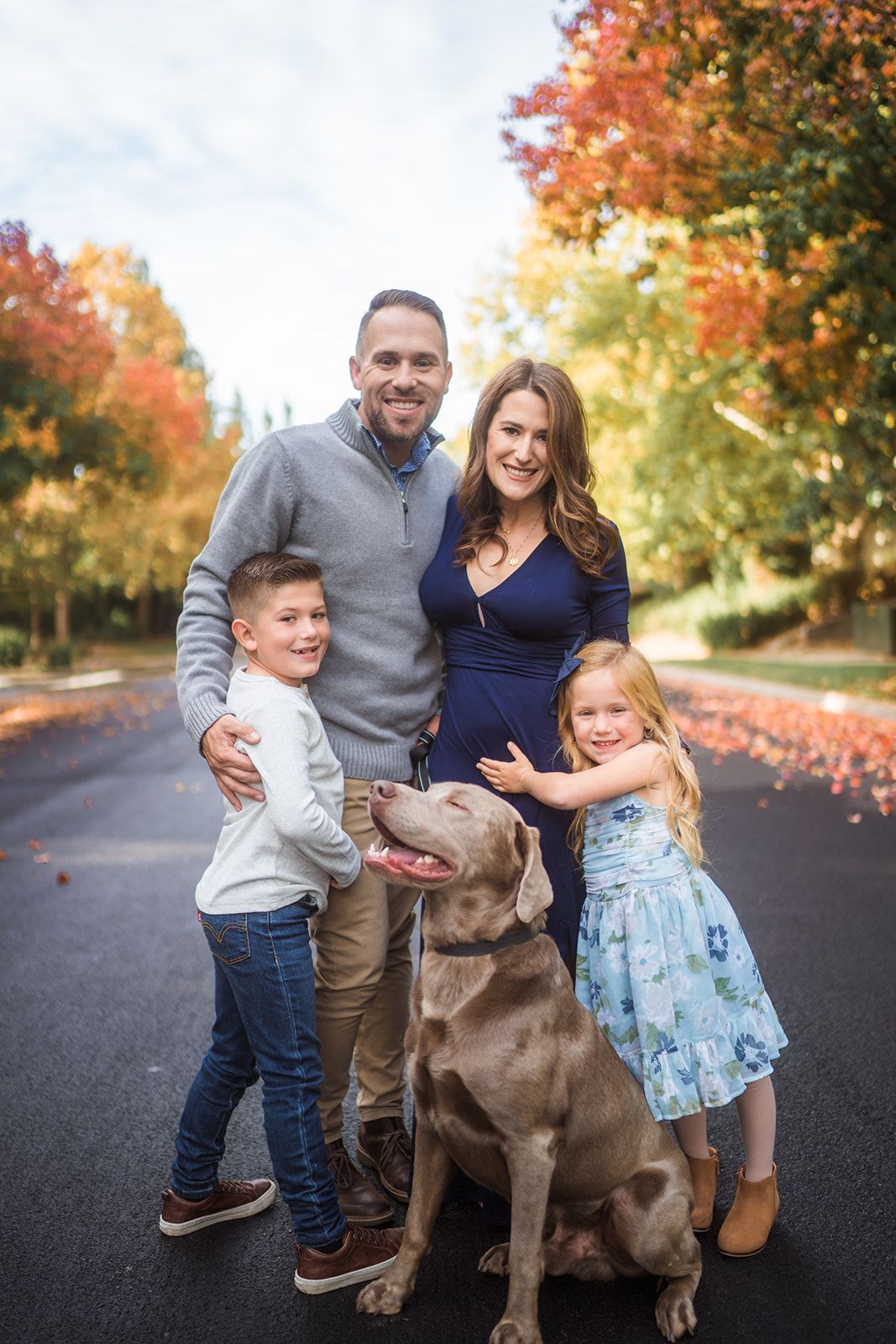 A family and their chocolate labrador dog pose together on a fall day with colorful autumn leaves.