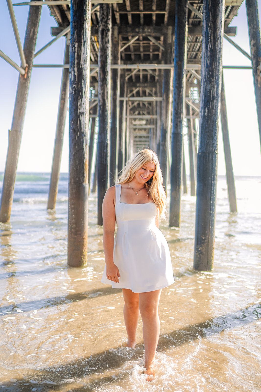 Blonde woman in white dress standing under a wooden beach pier with waves washing over her feet on a sunny day.