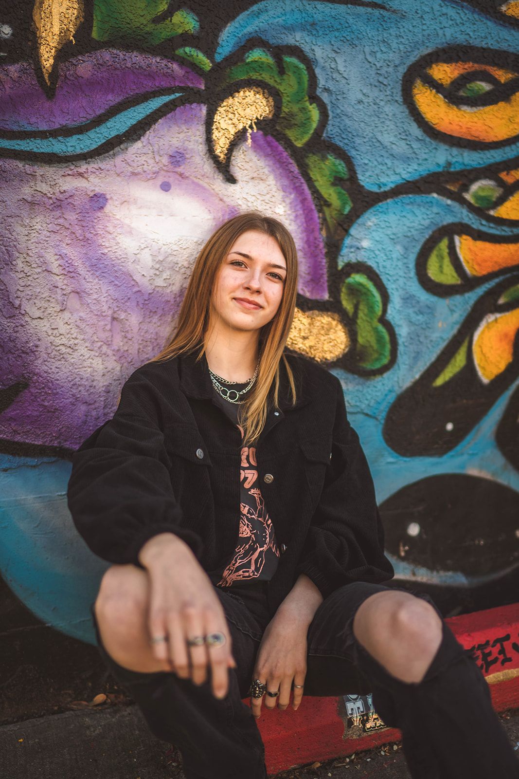 Young woman in black jacket sitting against colorful graffiti wall, wearing rings and smiling confidently.