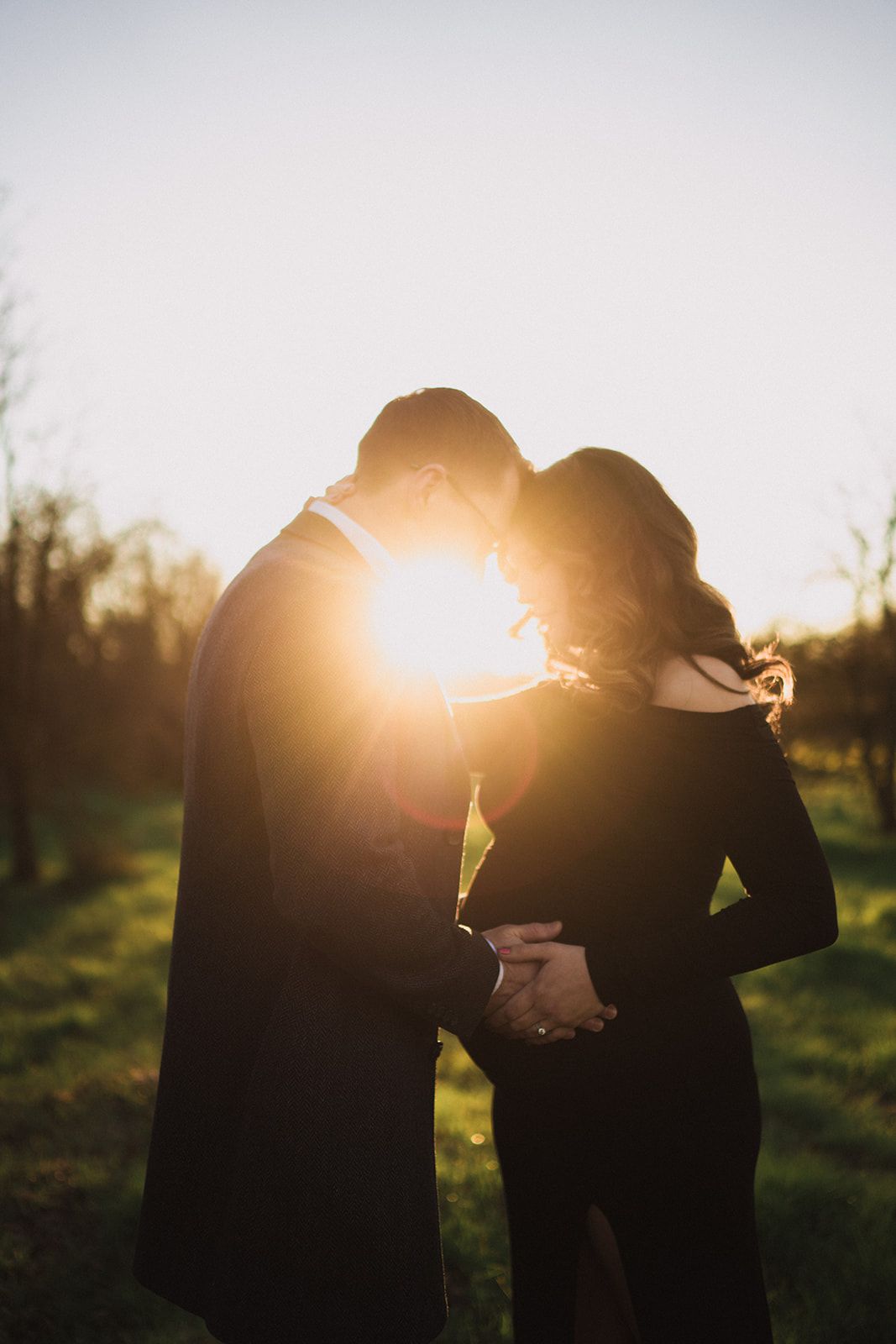 Couple in black embracing at golden sunset in a green park, backlit with warm glowing light.