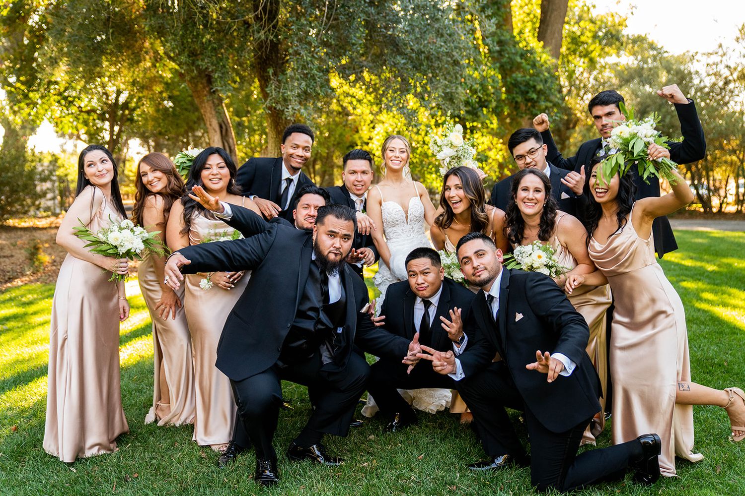 Wedding party in formal attire posing together on a sunny lawn with trees in the background.