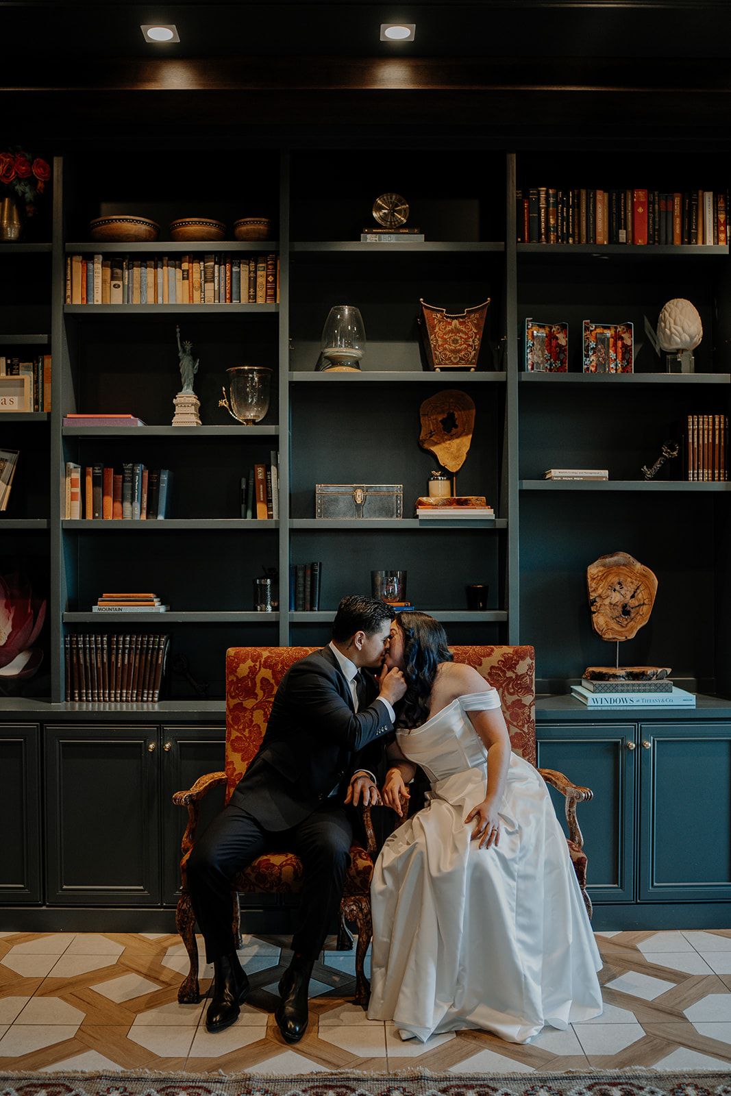 Bride and groom share a kiss while seated in ornate chairs before a dark teal bookshelf in an elegant library setting.