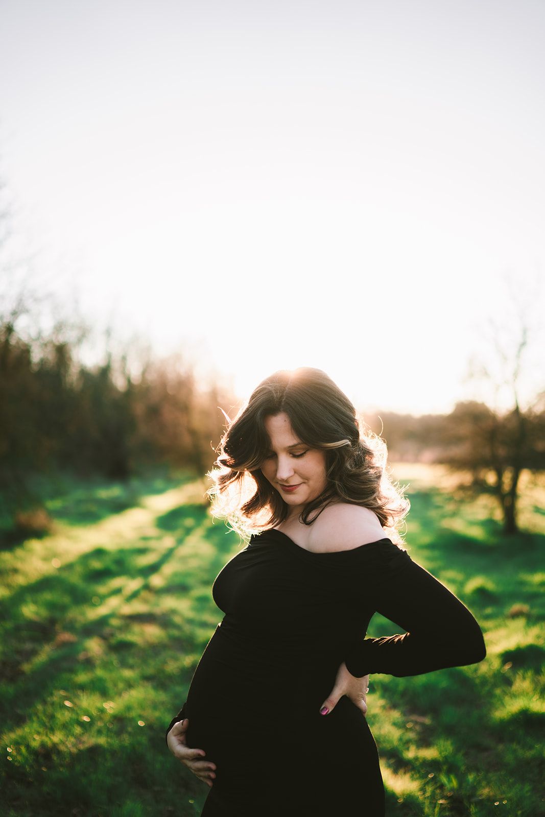 Pregnant woman in black off-shoulder dress holding baby bump outdoors at golden hour sunset.