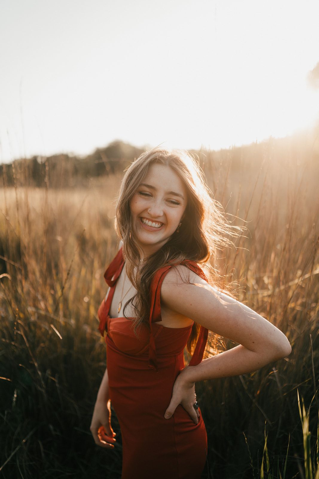 Young woman in red dress smiling in golden sunlit field with tall grass at sunset.