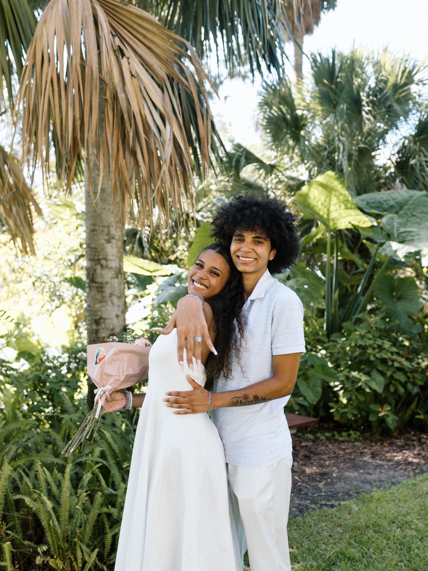 A couple embraces in a tropical garden setting with palm trees and lush foliage.