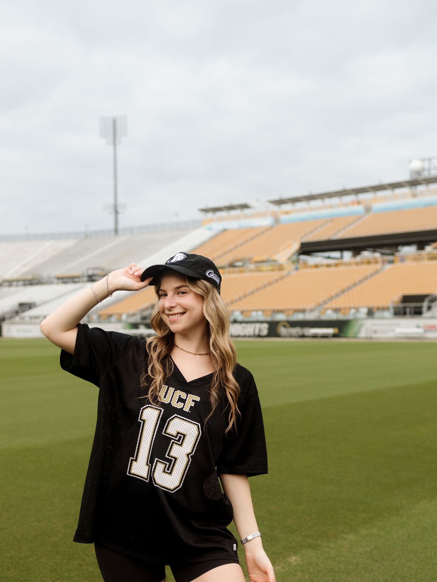 A person wearing a black UCF #13 jersey stands smiling on a football field with stadium seats in the background.