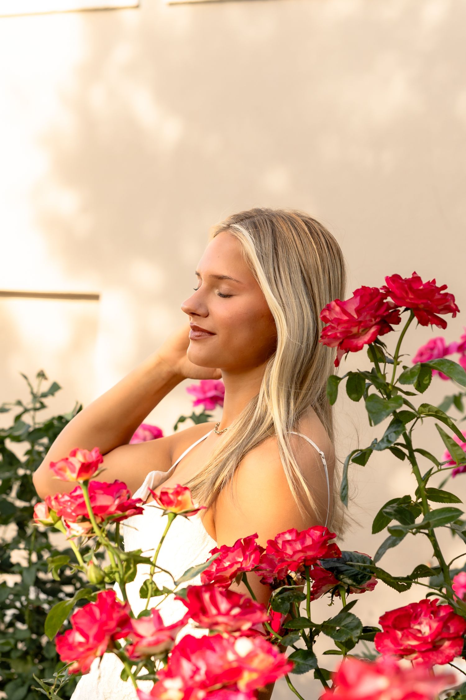 Blonde woman with eyes closed smelling red and pink roses in a sunlit garden.