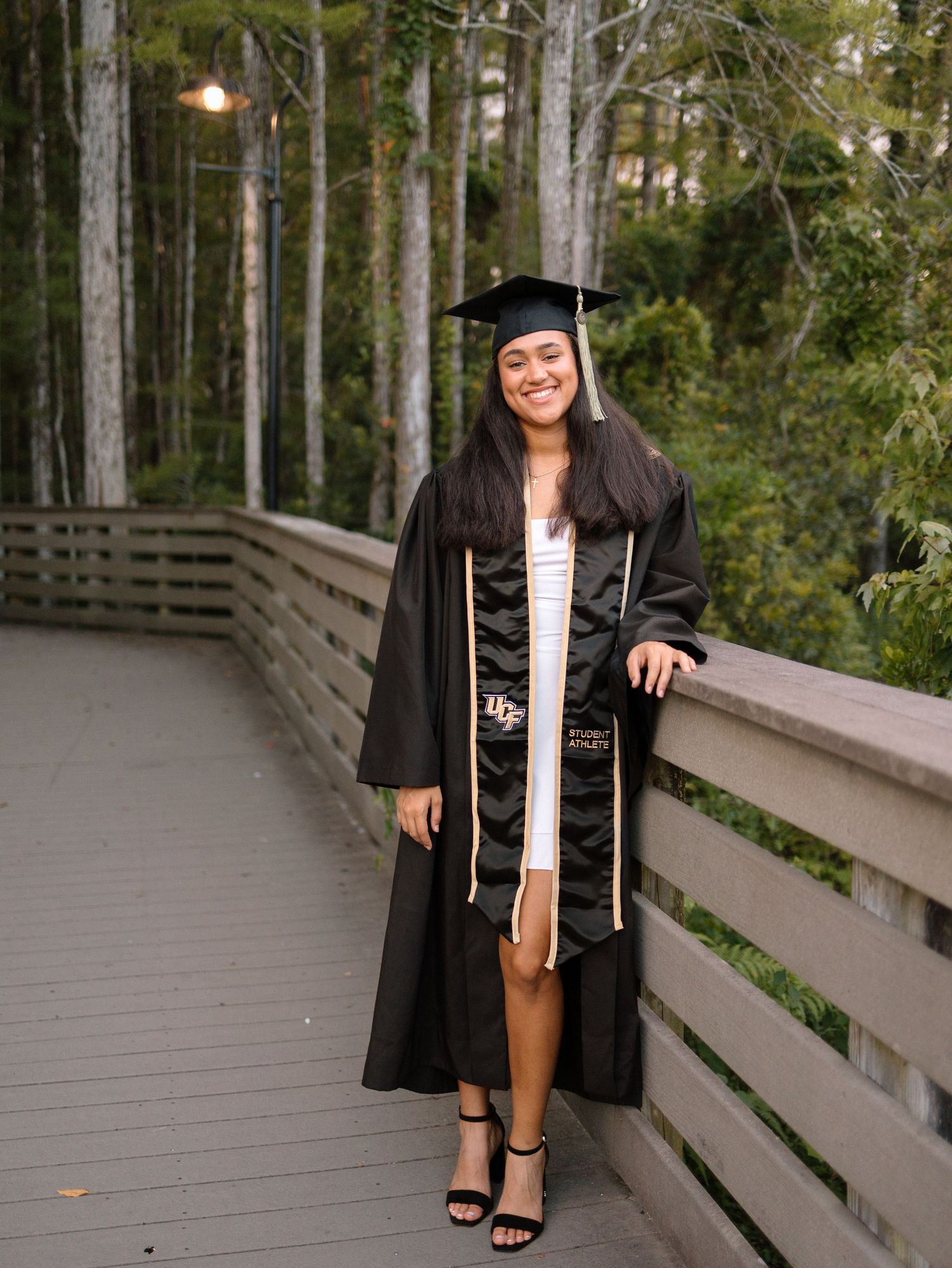 Smiling graduate in black cap and gown with gold stole poses on wooden bridge surrounded by trees.