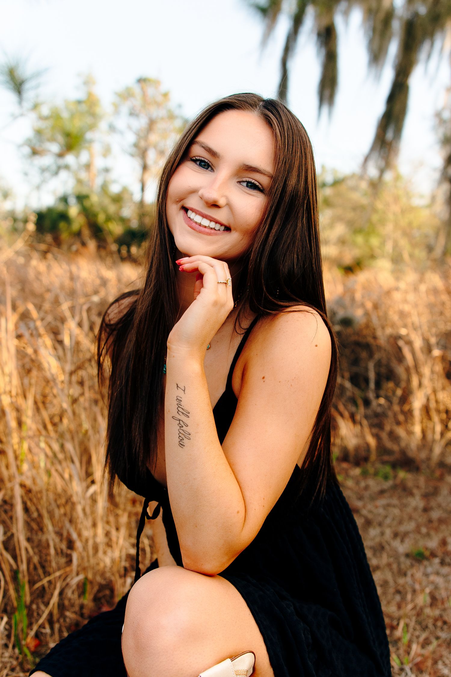 Smiling young woman with long dark hair and a forearm tattoo, wearing a black dress, posing outdoors in a golden field.