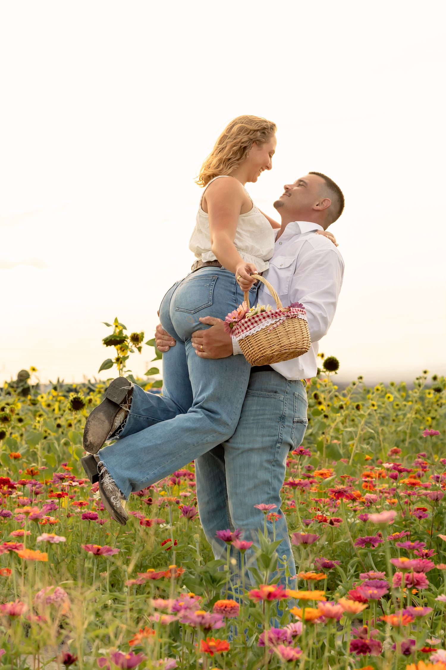 Couple embracing joyfully in a colorful flower field with sunflowers, man lifting woman while holding a picnic basket.