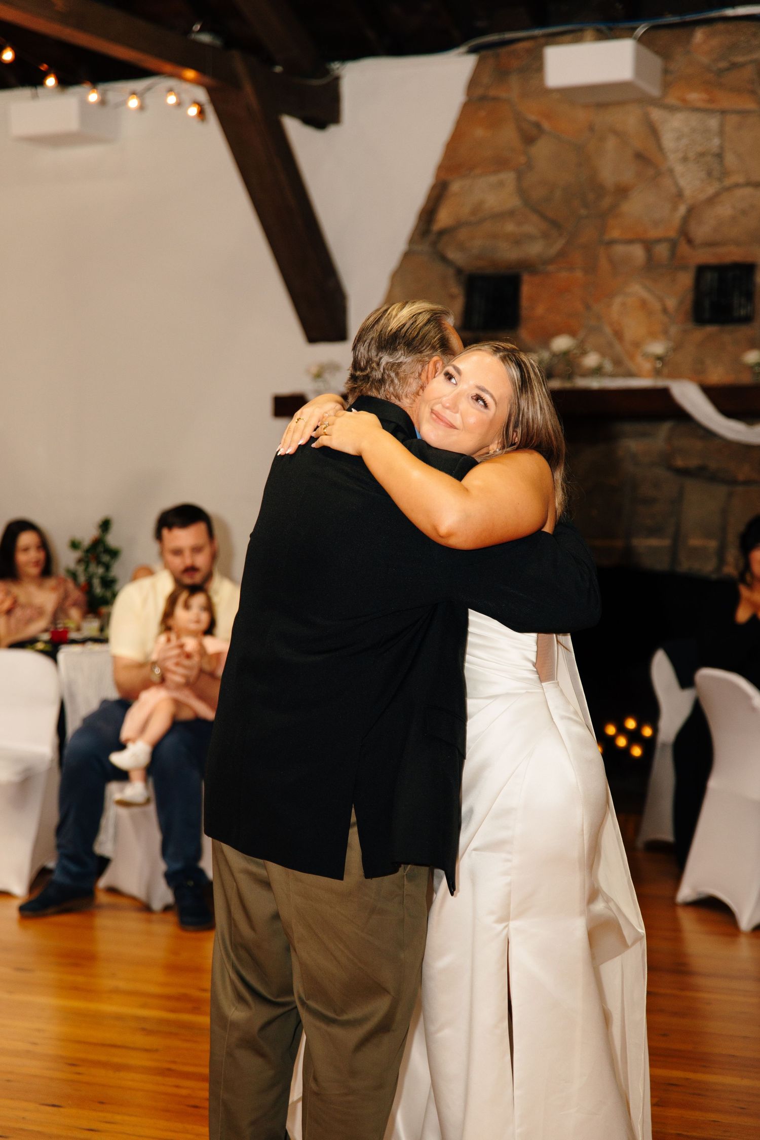 Bride in white gown shares emotional first dance hug with father at rustic wedding reception venue.