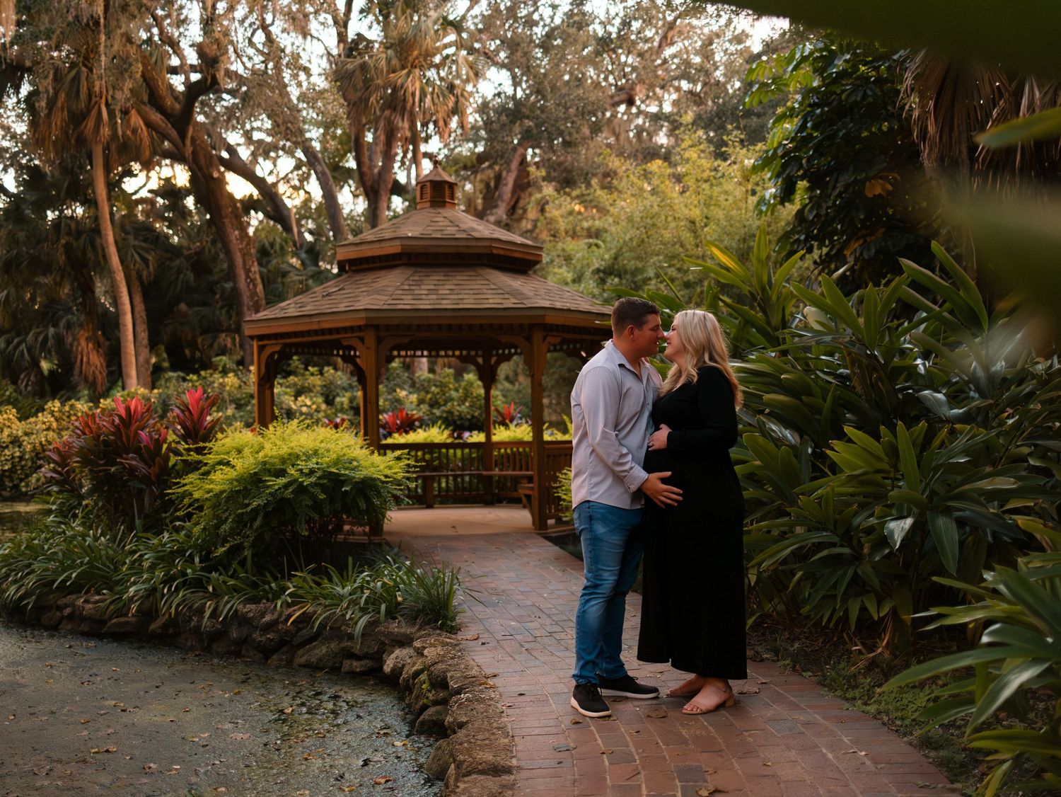 Couple sharing a tender moment in a lush garden with a wooden gazebo, woman in black dress cradling baby bump.