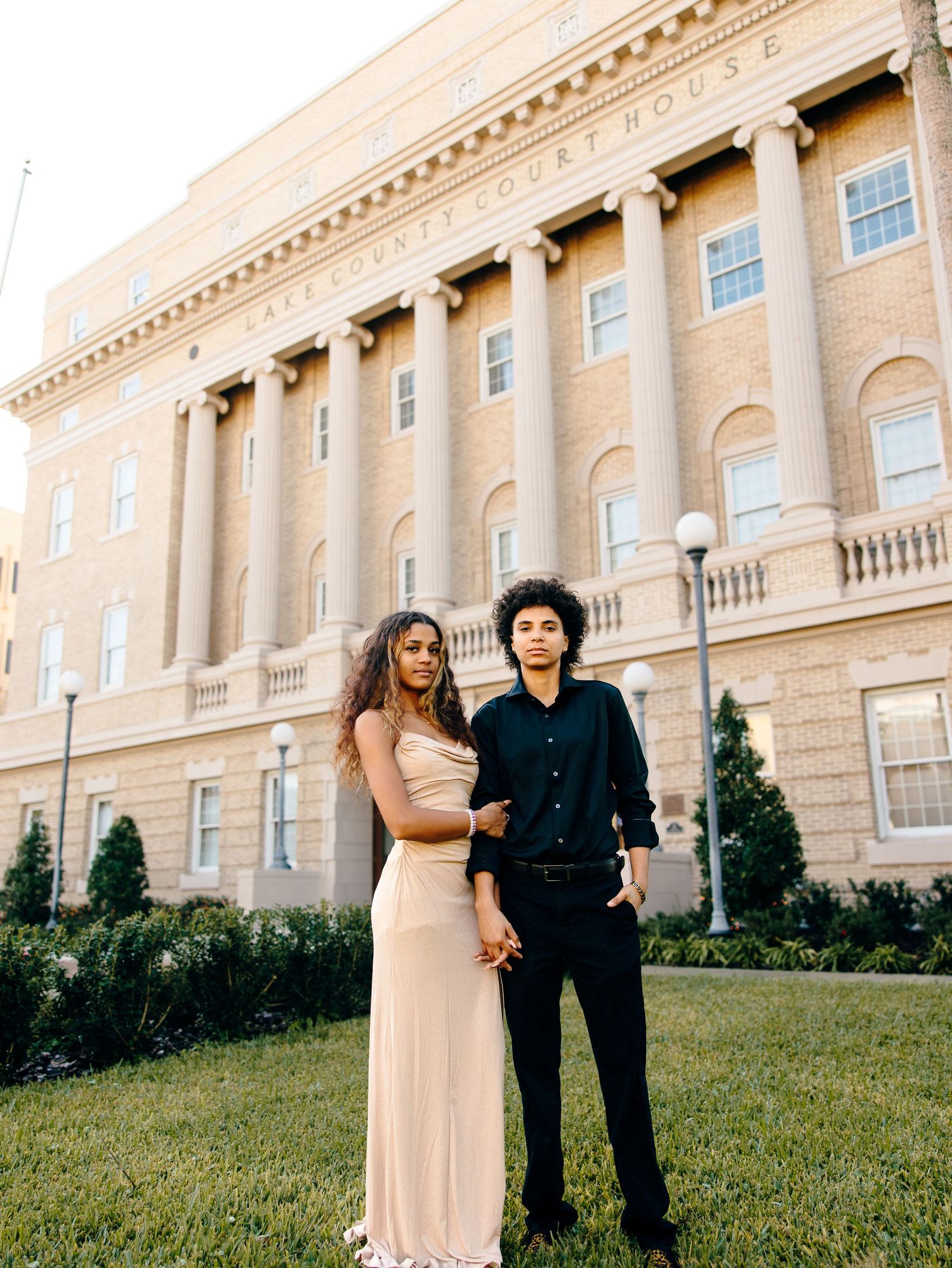 A couple poses in formal attire in front of a county courthouse, the woman in a cream gown and man in black.