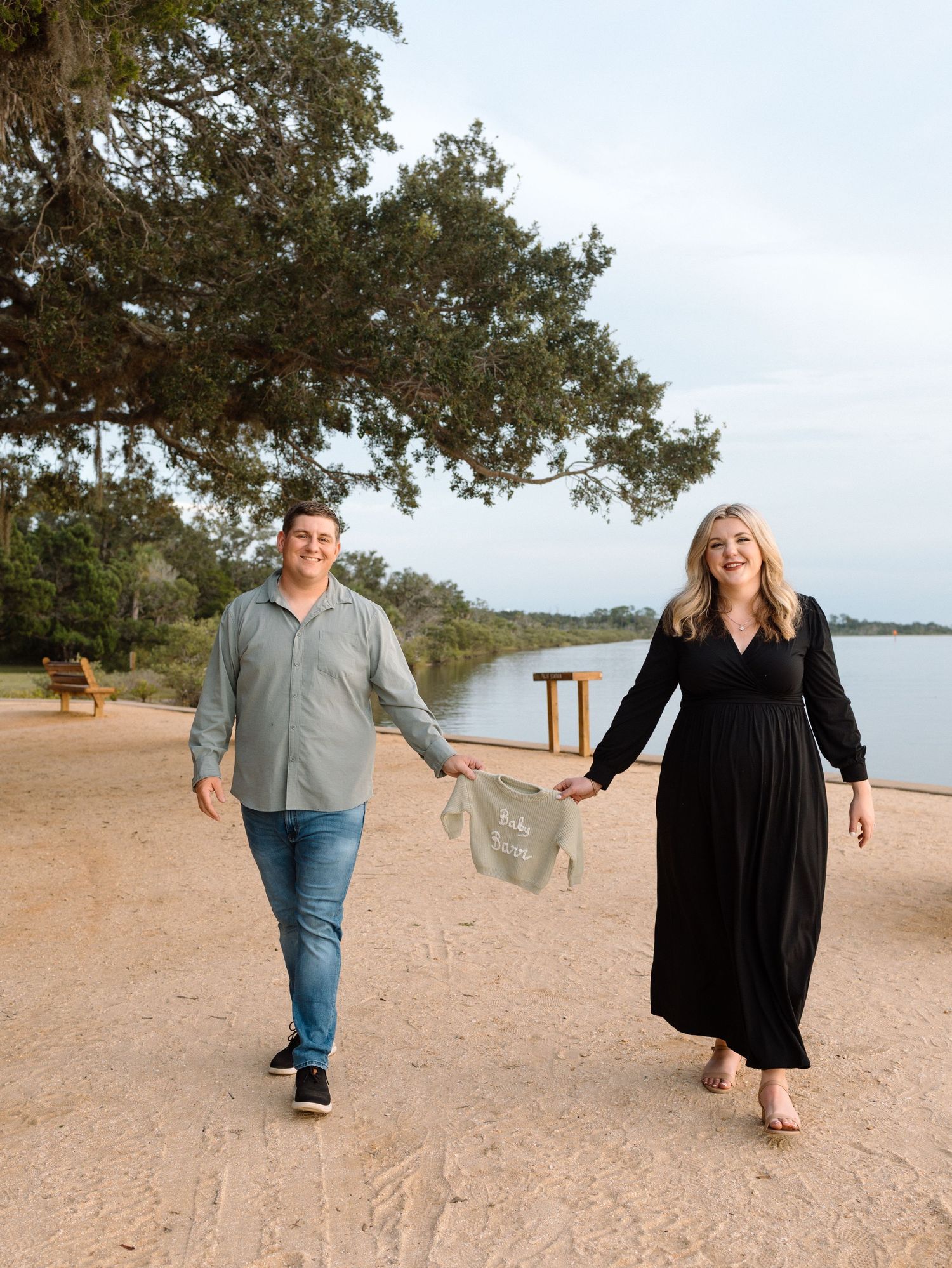 Couple holding baby onesie while walking on sandy beach near lake, maternity announcement photo session outdoors.