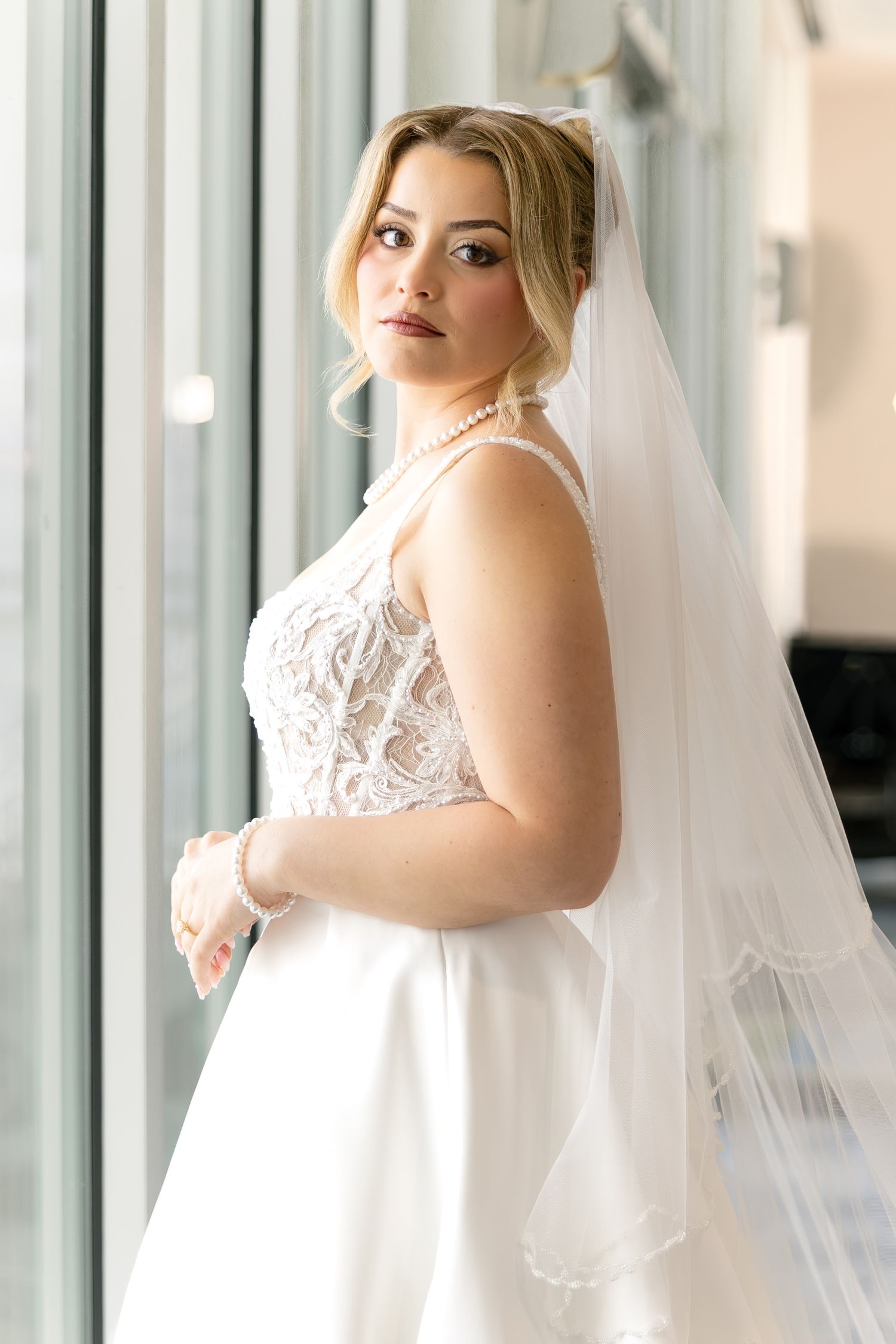 Elegant bride in white lace wedding dress and veil poses by window with pearl necklace and updo hairstyle.