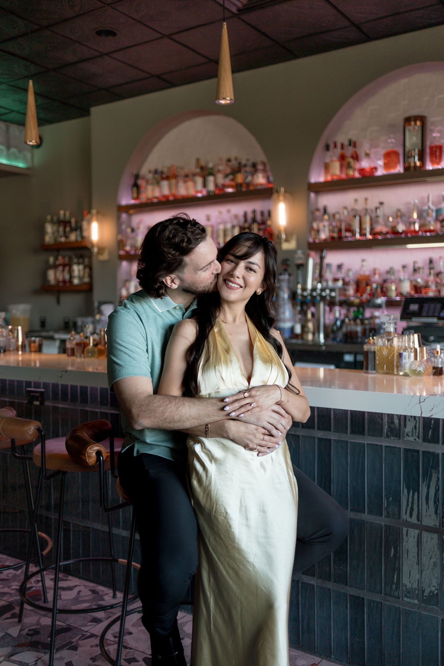A couple shares an intimate moment together at an elegant bar with bottles displayed on illuminated shelves behind them.
